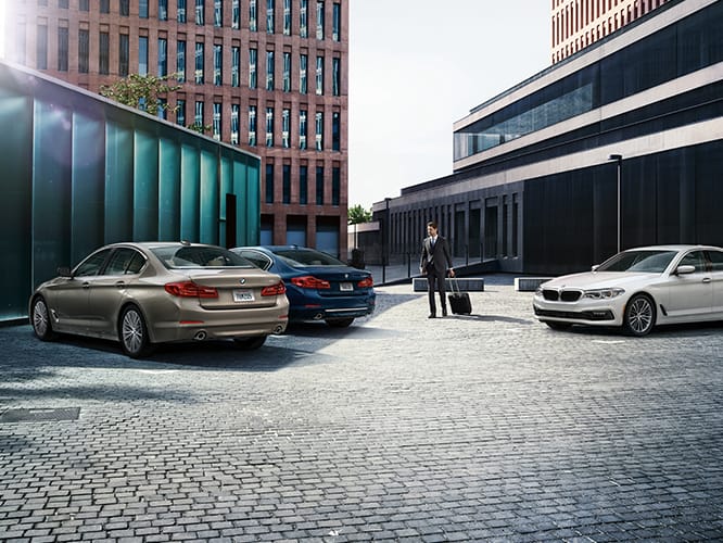 A business traveler with luggage approaches two parked BMW Sedans