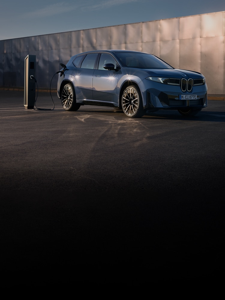 The 2027 BMW iX3 charging at a public charging station in front of a shimmering silver wall.